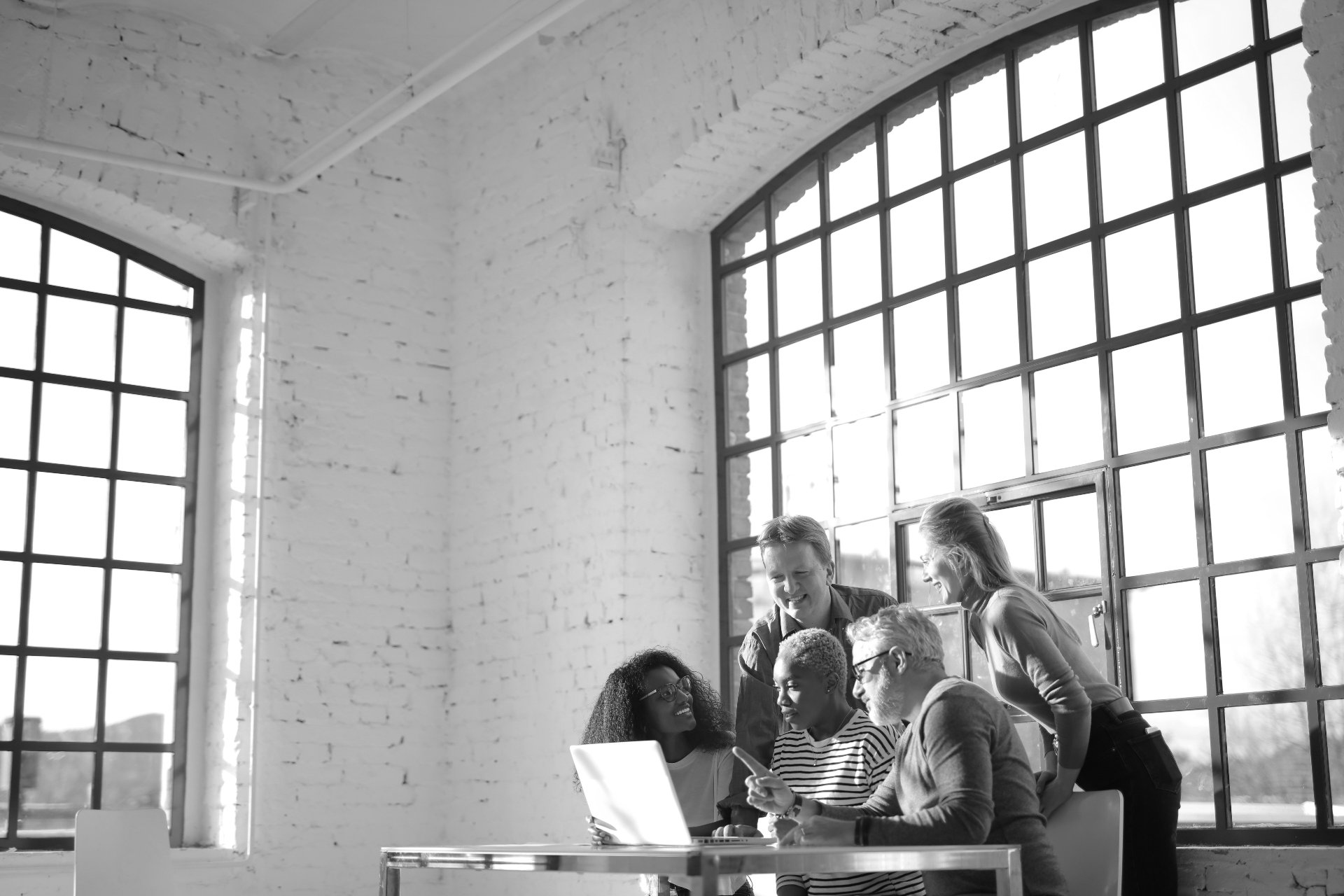 Five colleagues gathered around a laptop in a bright loft office with exposed brick walls and large industrial windows
