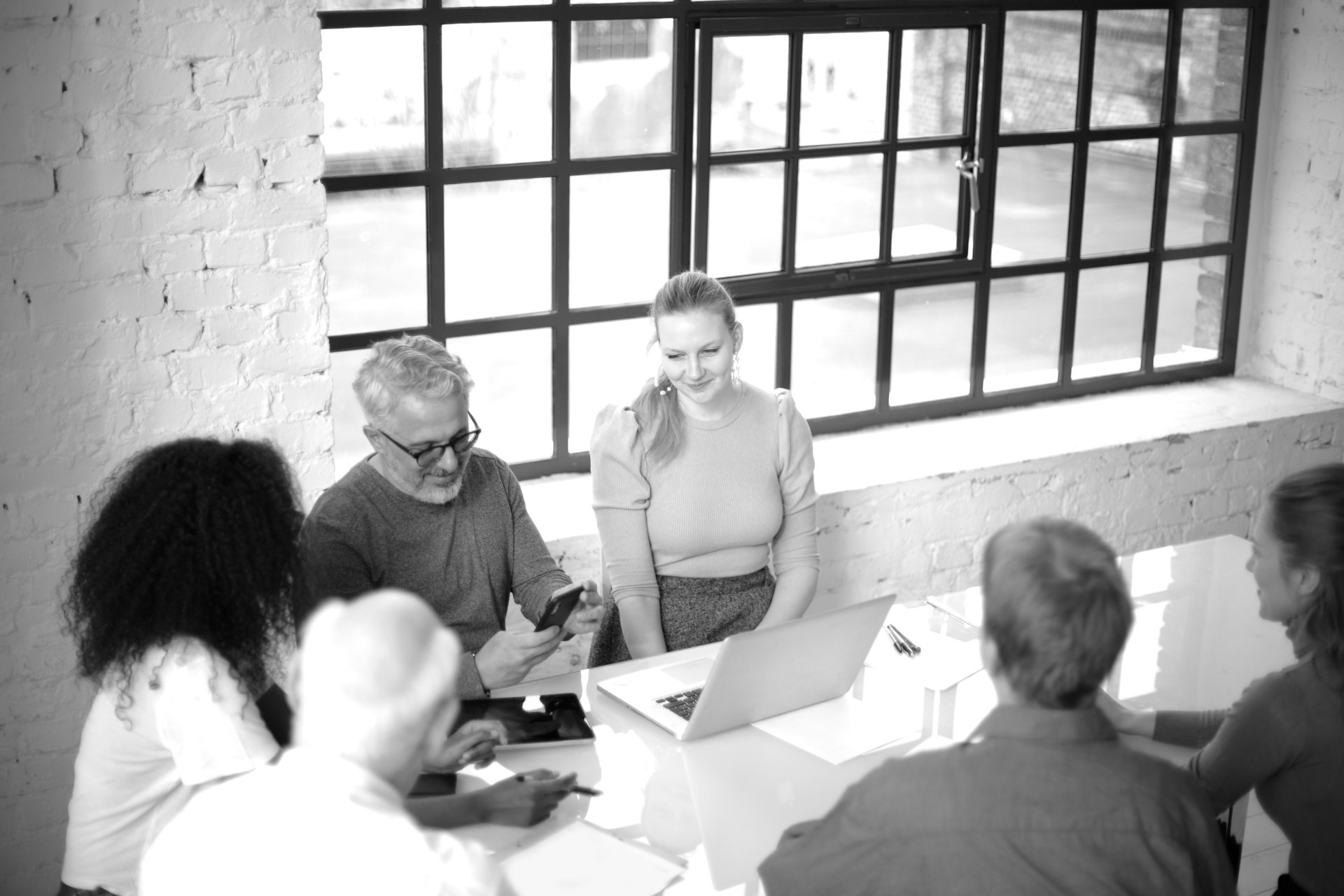 Six colleagues gathered around a laptop and tablet, reviewing work together in a bright, brick-walled loft office.