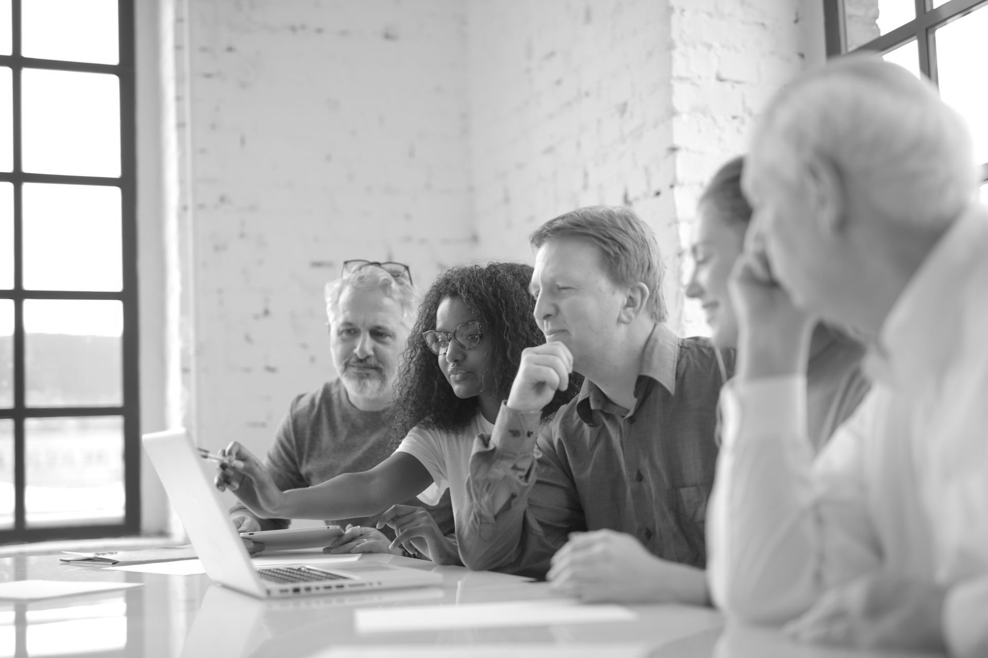 Five colleagues gathered around a laptop at a bright, brick-walled office table, one pointing at the screen.