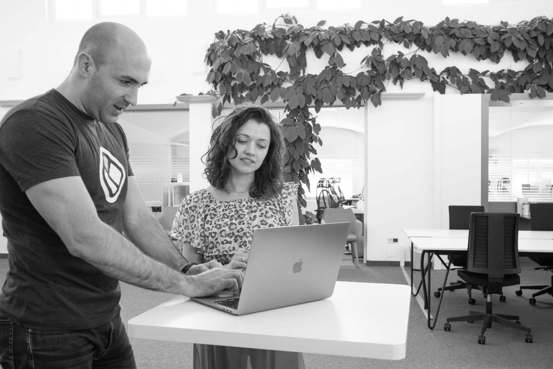 Two colleagues standing together at a laptop in a bright, plant-filled office, one pointing at the screen.
