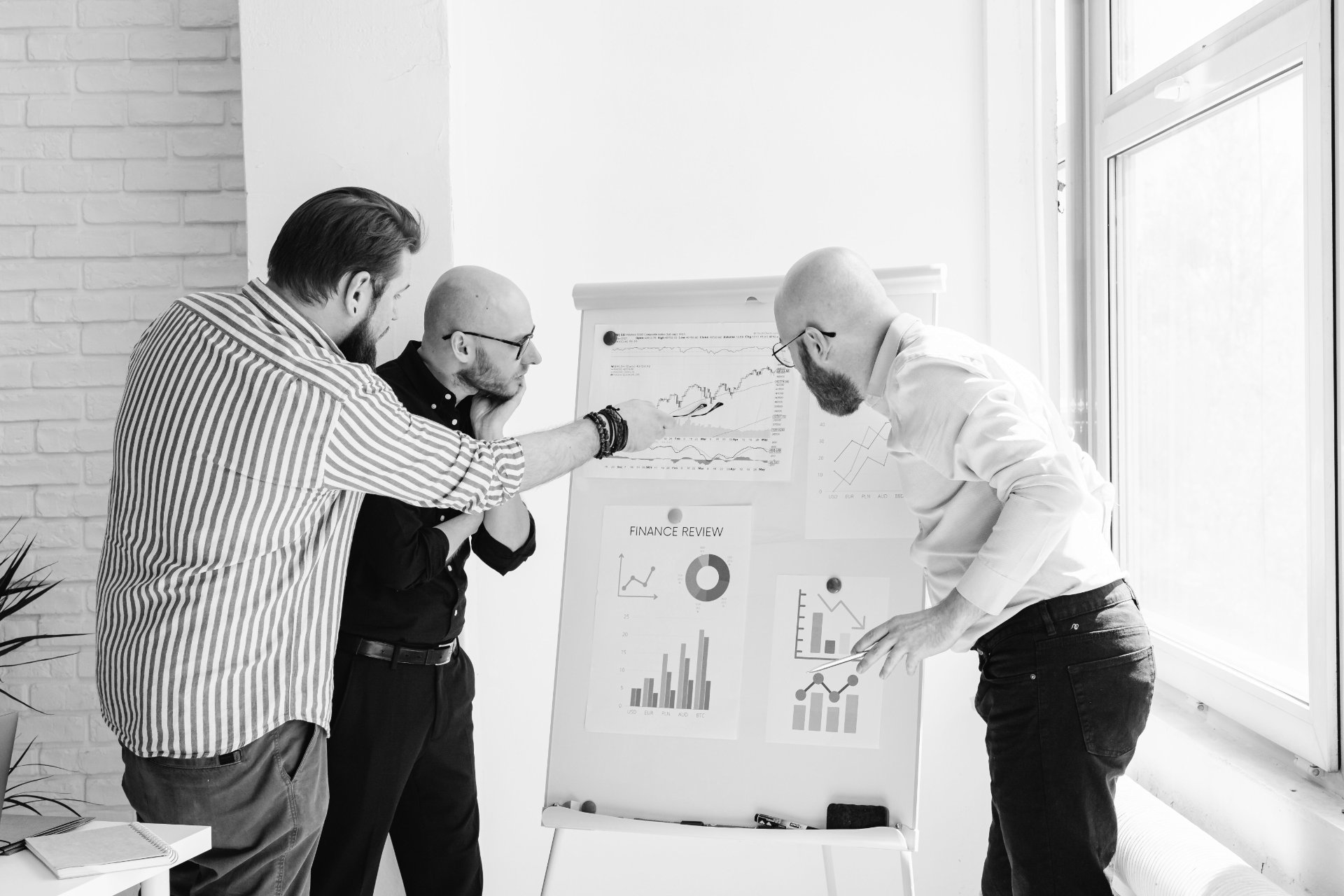 Three colleagues reviewing financial charts on a whiteboard in a brick-walled office